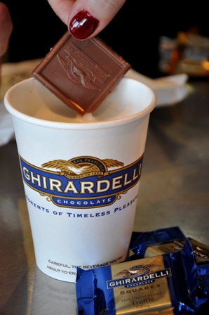 An image of Ghirardelli chocolate squares dipping into hot chocolate in a cup